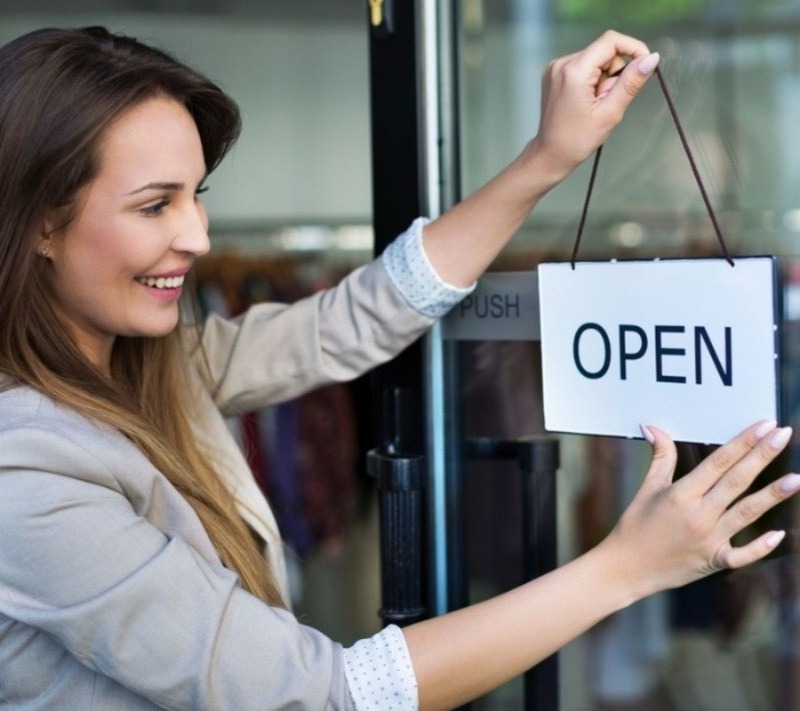 A picture of a smiling woman with a suit on. She is putting an open sign up on a door.